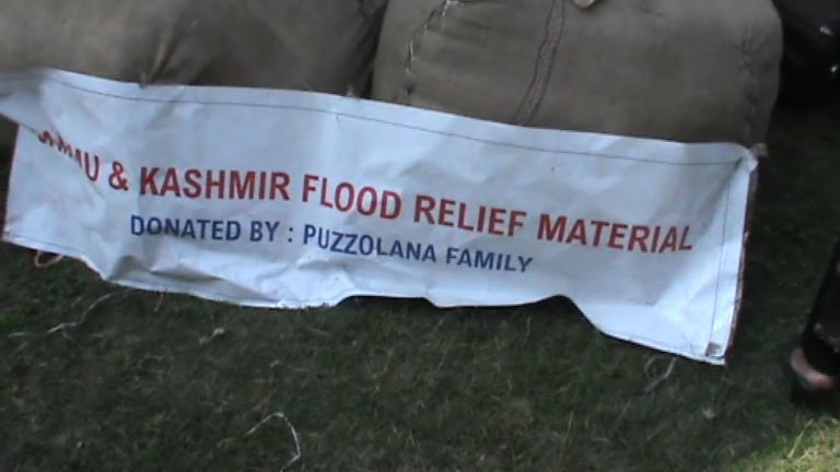 JK Flood Relief' against a backdrop of flood-affected areas, showing people receiving aid, emergency supplies being distributed, and volunteers working on relief efforts.